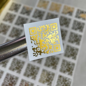 May include: A gold-colored QR code sticker on a white background. The sticker is surrounded by other blurred QR code stickers.