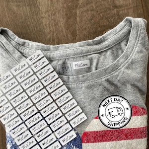 May include: A heather gray Gap Kids t-shirt featuring a sequined American flag design on the front. A sheet of rectangular white adhesive labels with the name McCann printed in black script is placed over the shirt, with one label applied to the collar.