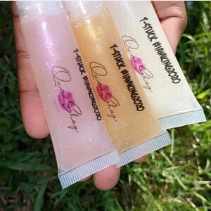May include: Three clear lip gloss tubes with a glittery texture. Each tube has a white label with the text "T-STUCK #IAMKING2020" and "Quo Shay" printed on it.