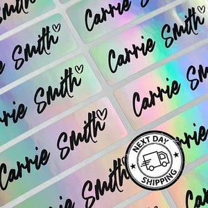 May include: A sheet of rectangular adhesive labels with a holographic, iridescent finish. Each label features the name Carrie Smith printed in a bold black script font, accompanied by a small heart icon. The labels are arranged in a grid pattern.