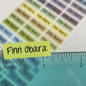 May include: A sheet of rectangular adhesive labels in various pastel colors, each printed with the name Finn Obara in black text. A teal ruler is positioned along the bottom edge, showing the label width is approximately 1.25 inches.