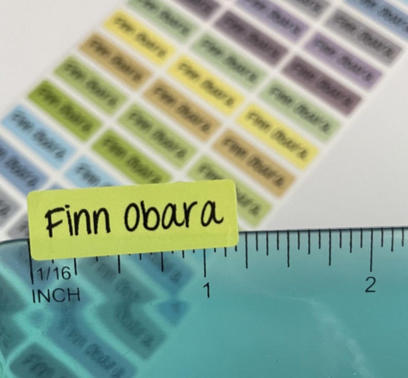 May include: A sheet of rectangular adhesive labels in various pastel colors, each printed with the name Finn Obara in black text. A teal ruler is positioned along the bottom edge, showing the label width is approximately 1.25 inches.