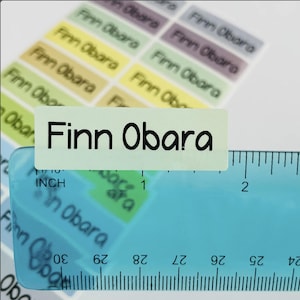 May include: A sheet of rectangular adhesive labels in various pastel colors, each printed with the name Finn Obara in a black sans-serif font. A blue plastic ruler is placed over the labels, showing a width of approximately 1.75 inches.
