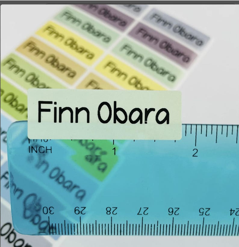 May include: A sheet of rectangular adhesive labels in various pastel colors, each printed with the name Finn Obara in a black sans-serif font. A blue plastic ruler is placed over the labels, showing a width of approximately 1.75 inches.