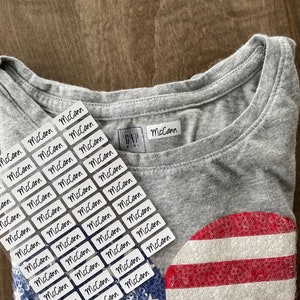May include: A sheet of white rectangular labels with the name "McCann" written in black cursive. The labels are placed on a gray t-shirt with a red, white, and blue sequined heart design. The t-shirt has a white tag with the text "GAP Kids" on it.