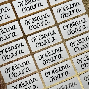 以下が含まれることがあります： 黒い筆記体で「Orellana Obara」と書かれた白いラベルが複数、金色の表面に並べて置かれている様子のクローズアップ。