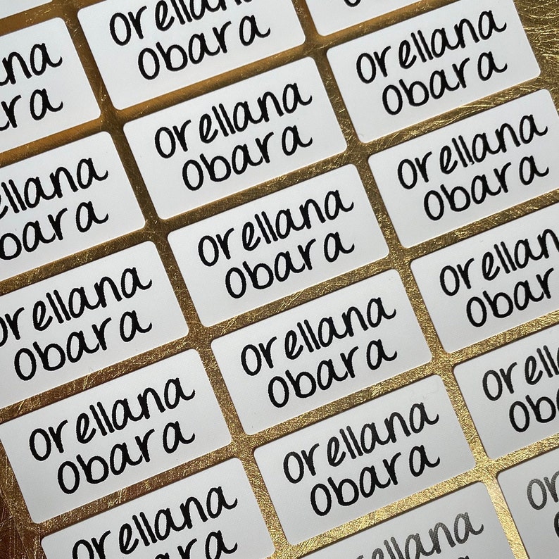 以下が含まれることがあります： 黒い筆記体で「Orellana Obara」と書かれた白いラベルが複数、金色の表面に並べて置かれている様子のクローズアップ。