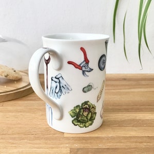 Op de afbeelding: Witte keramische mok met een handvat, met een kleurrijke illustratie van tuingereedschap, waaronder handschoenen, een troffel, een kruiwagen en een snoeischaar.