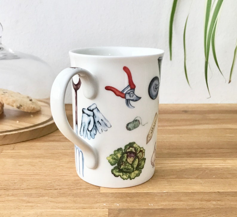 Op de afbeelding: Witte keramische mok met een handvat, met een kleurrijke illustratie van tuingereedschap, waaronder handschoenen, een troffel, een kruiwagen en een snoeischaar.