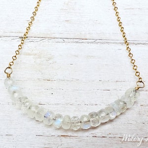 May include: A delicate gold chain necklace with a row of clear moonstone beads. The necklace is on a white wooden background.