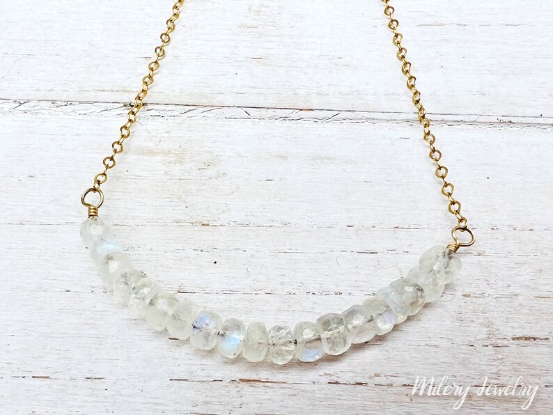 May include: A delicate gold chain necklace with a row of clear moonstone beads. The necklace is on a white wooden background.