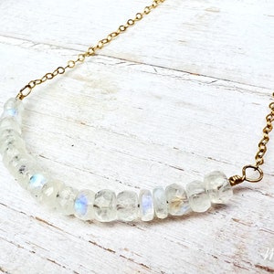 May include: A delicate gold chain necklace with a row of iridescent white moonstone beads. The necklace is on a white wooden surface.