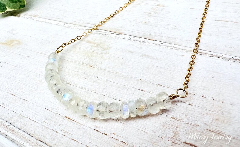 May include: A delicate gold chain necklace with a row of iridescent white moonstone beads. The necklace is on a white wooden surface.