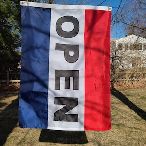May include: A vertical flag with blue, white, and red stripes, with the word "OPEN" in large black letters. The flag is hanging outside on a sunny day.