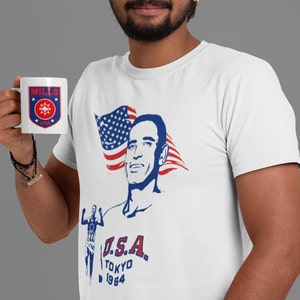 May include: White t-shirt with a blue and red American flag design. The flag is behind a portrait of a man with the text "U.S.A. TOKYO 1964" below. The man is wearing a blue shirt and has a determined expression on his face. The shirt also features a small logo with the text "MILLS" and a star design.