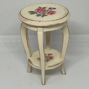 May include: A small, cream-coloured decorative plant stand with a round top and a lower shelf. The top and shelf feature a floral design with pink, red, and blue flowers. The stand has three curved legs and is painted with a distressed finish.