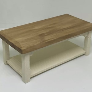 May include: A miniature wooden coffee table. The top is natural wood-tone, and the base and lower shelf are cream-coloured. The table has four legs and is designed for a doll's house or miniature setting.