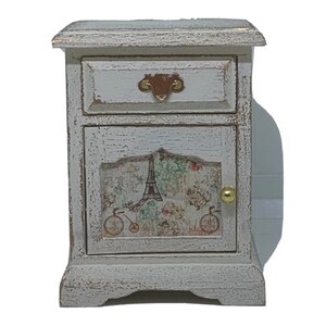 May include: A small white wooden bedside table with a single drawer and a door. The door has a printed design featuring the Eiffel Tower and bicycles. The bedside table has a distressed finish and gold accents.