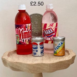 May include: Miniature soft drink bottles and cans on a small wooden table. The collection includes a red VIMTO bottle, a pink BARR Cream Soda bottle, and Pepsi and Fanta cans. The items are arranged with a coin in the background.