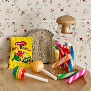 Puede incluir: Escena en miniatura con un tarro de cristal lleno de bastones de caramelo de colores, una bolsa amarilla de "Bassetts Jelly Babies", dos piruletas y una moneda. El tarro tiene un tapón de corcho. La escena está sobre una superficie de madera.