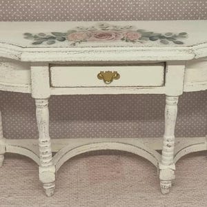 May include: A small, white, half-moon table with a distressed finish. The table has a single drawer with a gold-coloured handle and is decorated with a floral design in pink and green. The table is supported by four ornate legs.