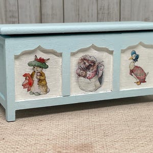 Puede incluir: Un cofre de madera azul claro con tres paneles que muestran ilustraciones de un conejo, un erizo y un pato. El cofre tiene una tapa con bisagras y está decorado con un fondo blanco.