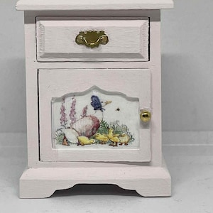 Puede incluir: Una mesita de noche rosa en miniatura con un cajón y una puerta de armario. La puerta del armario presenta una colorida ilustración de una madre pato y sus patitos. El cajón tiene un tirador dorado y la puerta tiene un pomo dorado.
