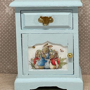 Puede incluir: Una mesita de noche de madera azul claro con un solo cajón y una puerta de armario. La puerta del armario tiene una imagen impresa de tres conejos con chaquetas rojas. El cajón tiene una manija dorada.