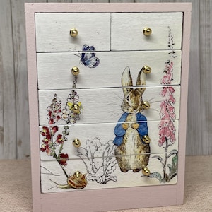 Può includere: Un piccolo baule di legno rosa con cinque cassetti. I cassetti sono decorati con un'illustrazione di Peter Rabbit che presenta un coniglio, una farfalla, fiori e una lumaca. Ogni cassetto ha una manopola dorata.