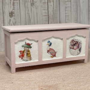 Puede incluir: Un cofre de madera rosa con tres paneles que muestran ilustraciones de un conejo, un pato y un erizo. El cofre tiene una tapa con bisagras y está decorado con un fondo blanco y un borde negro.