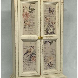 May include: A white painted wooden cabinet with two doors. The doors are decorated with floral and bird patterns and have gold knobs. The cabinet has a distressed finish and sits on four turned legs.