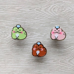 May include: Three enamel pins featuring cartoon frogs wearing blue hats. The frogs are green, pink, and brown.
