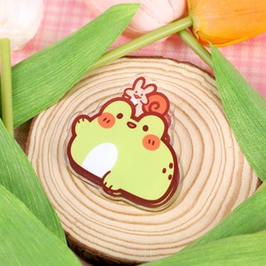 May include: A green frog with a brown snail on its head, both are cartoon characters. The frog is smiling and has a white belly. The snail is also smiling and has a brown shell. The frog and snail are on a wooden circle with a light brown background. The circle is surrounded by green leaves and pink tulips.