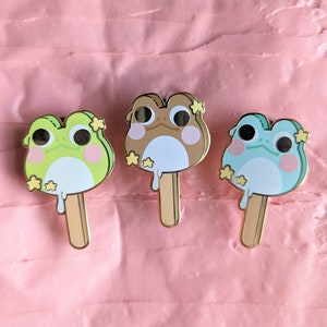 May include: Three enamel pins shaped like frog-faced popsicles. The pins are green, brown, and blue, with white drips and gold accents. Each frog has a star-shaped flower on its head and a gold stick.
