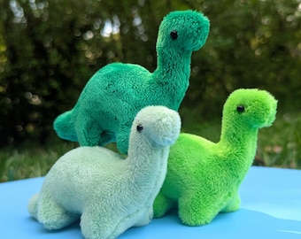 May include: Three plush green dinosaur toys. The dinosaurs are different shades of green and are arranged in a row on a blue surface.