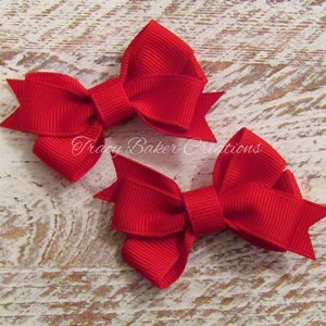 May include: Two bright red hair bows made of ribbed grosgrain ribbon. Each bow features a central knot and two pointed tails on either side. The text Tracy Baker Creations is printed in white script across the center of the image.