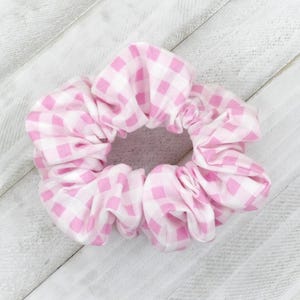 May include: A pink and white gingham scrunchie, a fabric hair accessory. The scrunchie is made of a soft, gathered fabric with a classic checkered pattern. The color scheme is a light pink and white.