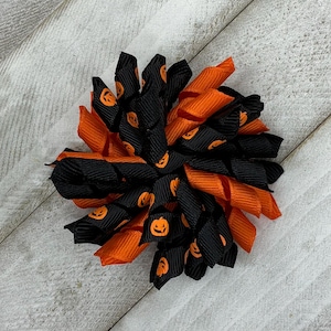May include: A circular hair accessory constructed from layered loops of grosgrain ribbon. The design features alternating black ribbons printed with small orange jack-o'-lantern faces and solid orange ribbons, arranged in a dense, ruffled flower shape.