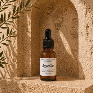 Puede incluir: Una botella de vidrio marrón con un cuentagotas negro, etiquetada como "Aegean Sun". La etiqueta enumera los ingredientes y el volumen como 15 ml. La botella se coloca sobre un fondo beige texturizado con una ramita verde.