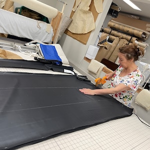 May include: A person cuts a large piece of black fabric with a rotary cutter on a gridded cutting mat. The fabric is marked with white lines.