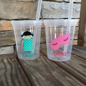 May include: Two clear plastic cups with straws, personalized with the name "Della". One cup has a girl in a sleeping bag with polka dots, the other has a pink sleep mask.