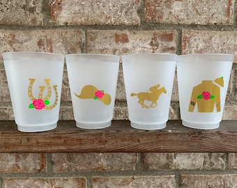 Puede incluir: Cuatro vasos de plástico transparente con diseños dorados y rosados. Los diseños incluyen una herradura, un sombrero, un caballo y un jinete, y una chaquetilla de jockey.
