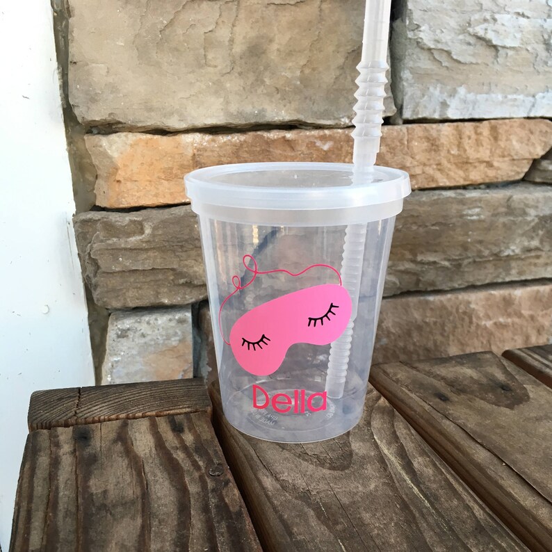 May include: A clear plastic cup with a pink sleep mask design and the name "Della" printed on the side. The cup has a straw and a lid.