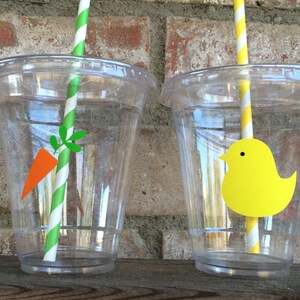 May include: Two clear plastic cups with straws and paper cutouts. One cup has a yellow chick cutout and the other has a carrot cutout.