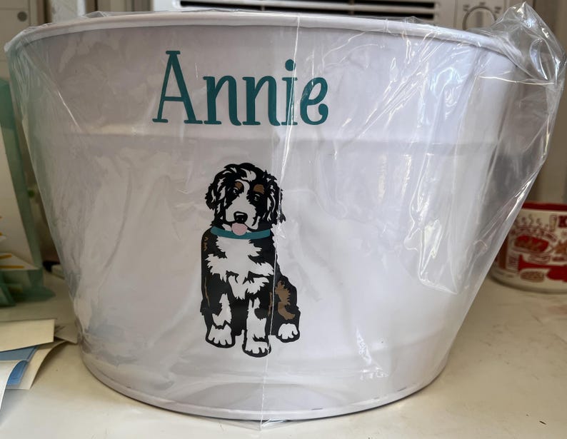 May include: White metal bucket with a black and white dog illustration and the name "Annie" printed in teal.