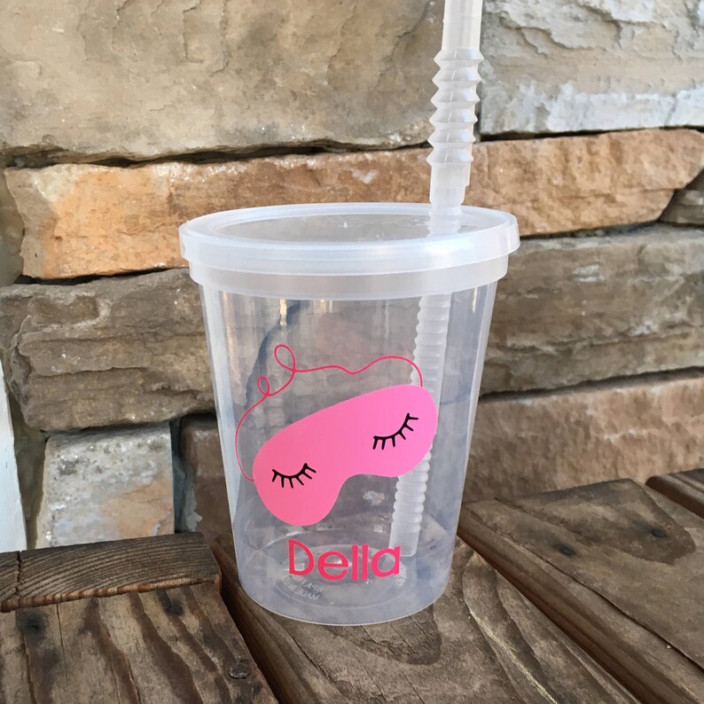 May include: A clear plastic cup with a pink sleep mask design and the name "Bella" printed on the bottom. The cup has a lid and a clear plastic straw.