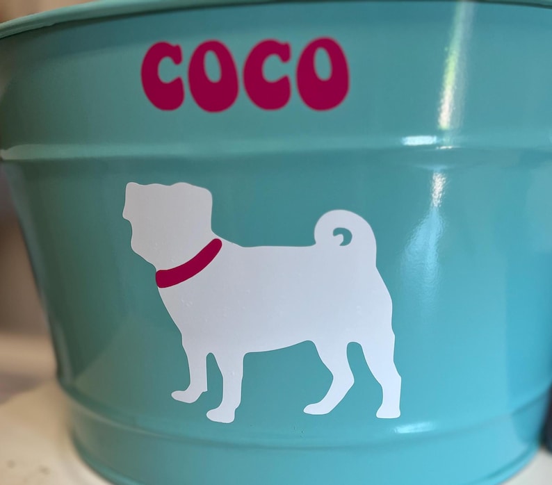 May include: A turquoise blue metal dog food bowl with a white silhouette of a pug and the name "Coco" in pink letters.