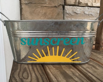 May include: A silver metal container with a yellow sun and the word "sunscreen" in teal lettering.