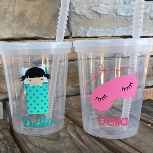 May include: Two clear plastic cups with lids and straws. The cups have a personalized design with the name "Della" and a sleep-themed image. One cup features a girl in a sleeping bag with polka dots, and the other cup features a pink sleep mask with eyelashes.