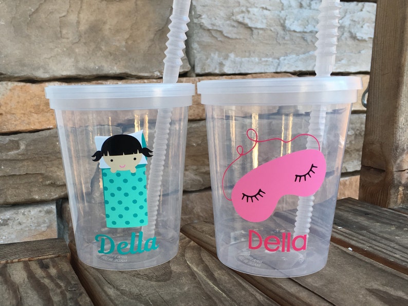 May include: Two clear plastic cups with lids and straws. The cups have a personalized design with the name "Della" and a sleep-themed image. One cup features a girl in a sleeping bag with polka dots, and the other cup features a pink sleep mask with eyelashes.